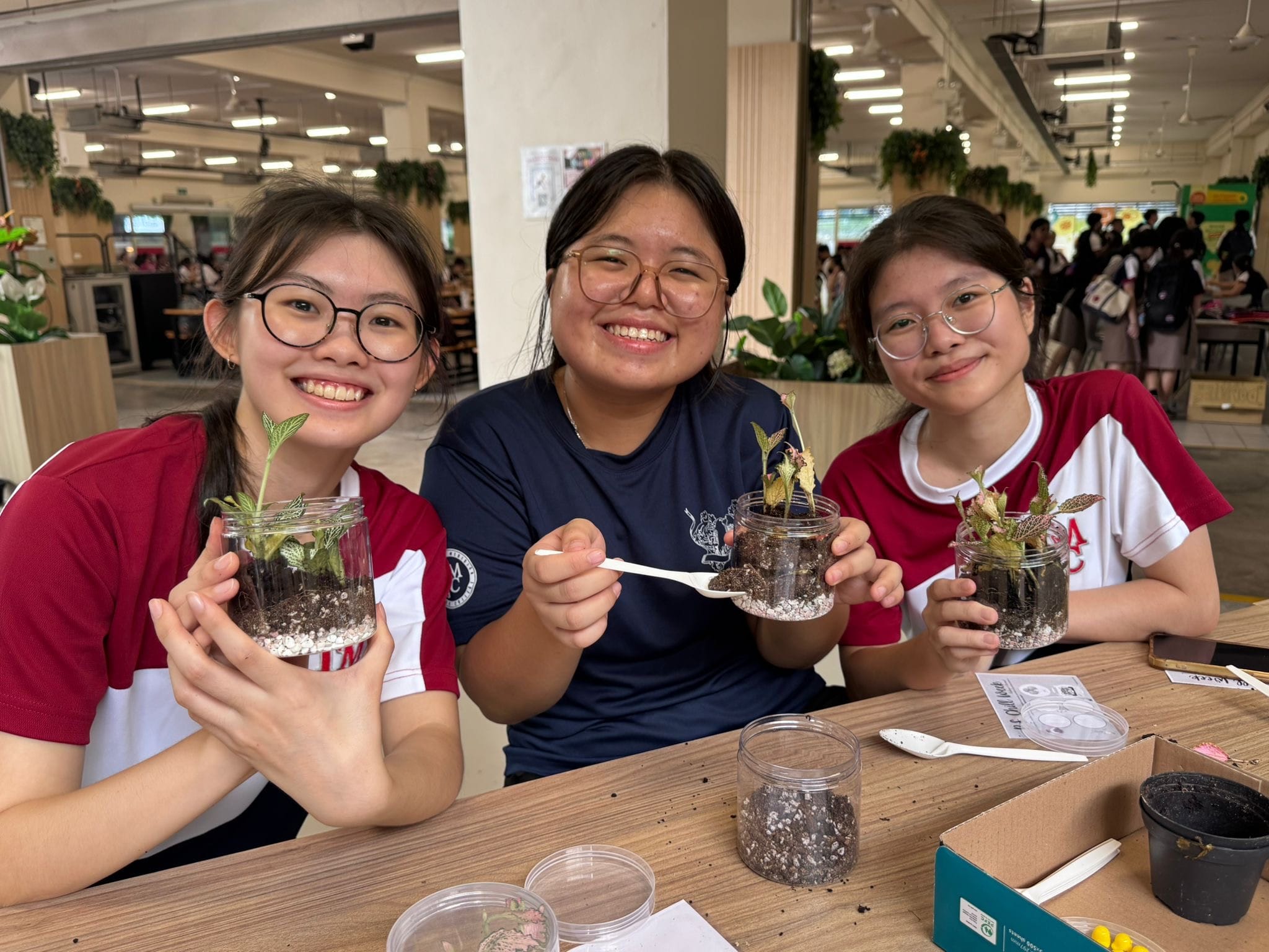 Three smiling female students in school uniforms hold up small glass jars containing soil, pebbles, and small plants — their handmade terrariums — at a canteen table.